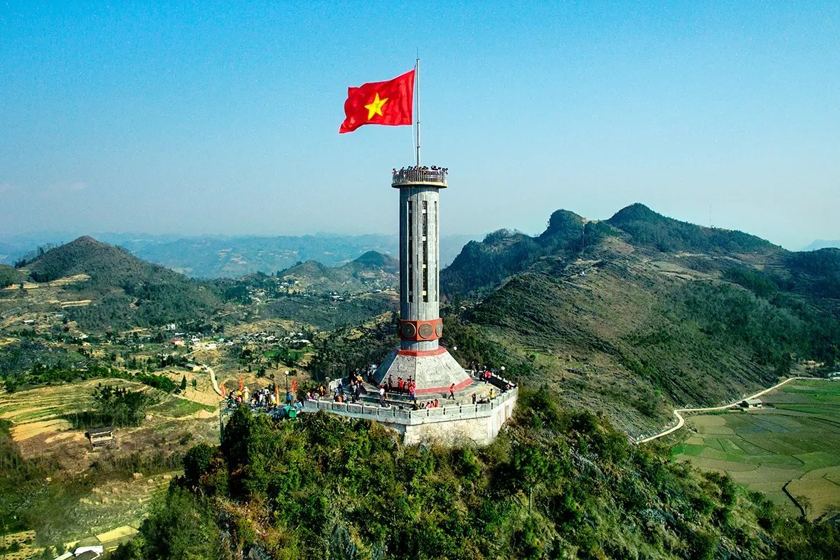 A close-up, detailed view of the North Vietnamese flag, the red flag with a yellow star, waving against a blurred Hanoi background, symbolizing the nation.