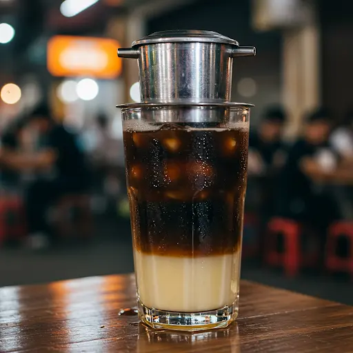 A tall glass showing distinct layers of dark iced coffee over sweetened condensed milk, topped with a Vietnamese phin drip filter, on a wooden table