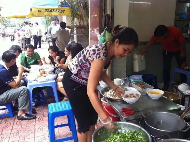 A woman prepares bowls of pho at a Vietnamese street food stall while customers eat seated on low plastic stools on the sidewalk. (1)
