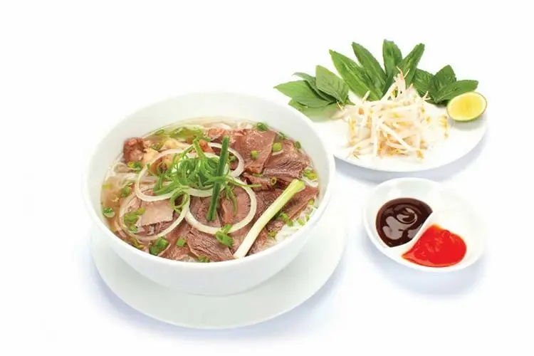 Bowl of Vietnamese beef Pho (Pho Bo) with side dishes of herbs, sprouts, lime, and sauces, isolated on a white background.