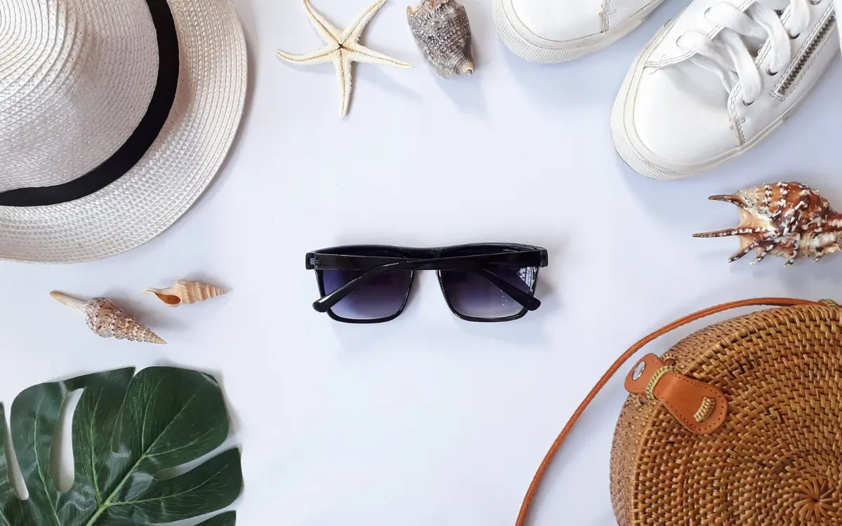 Flat lay of summer beach travel essentials including a straw hat, sunglasses, rattan bag, white sneakers, seashells, and starfish on a white background.