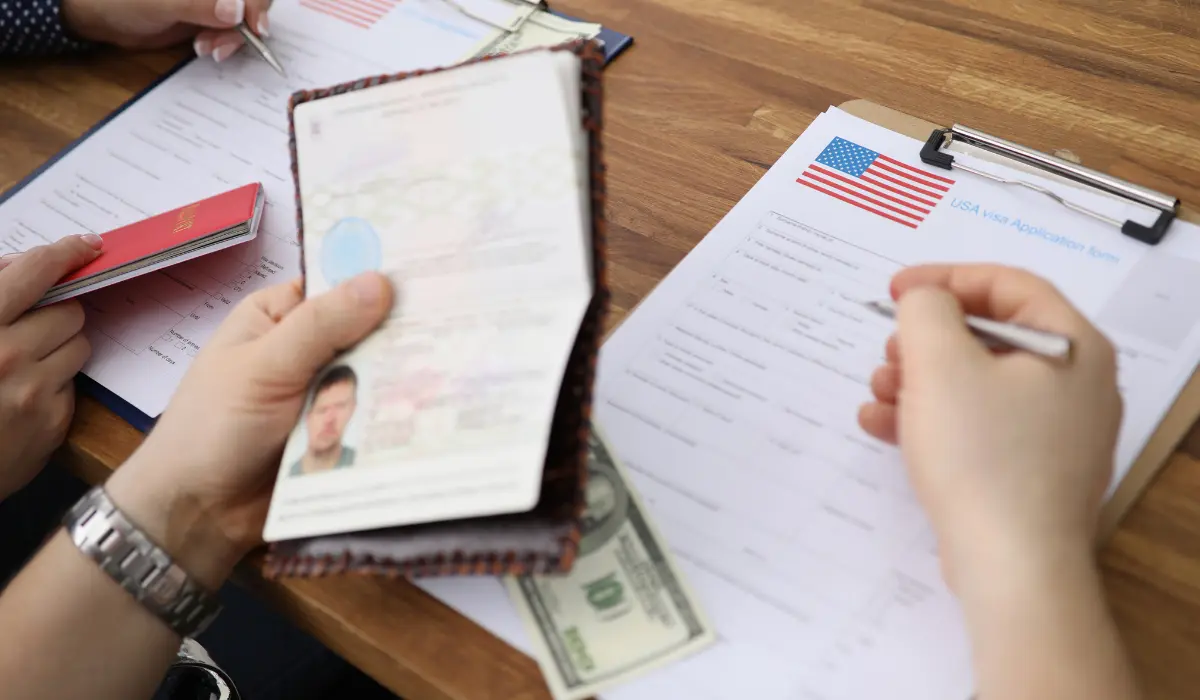 Hands filling out a visa application form next to an open passport on a desk.