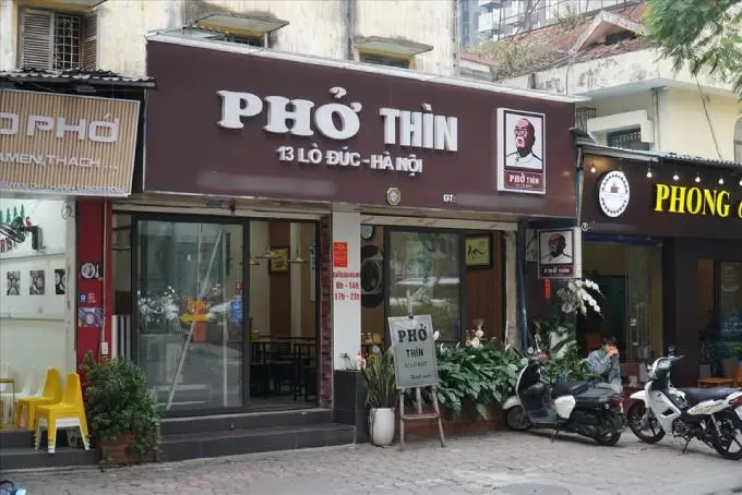 Photo of the storefront of the famous Pho Thin restaurant at 13 Lo Duc, Hanoi, with its distinctive brown and white sign, and motorbikes parked out front.