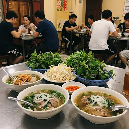 Three steaming bowls of Pho noodle soup with abundant fresh herbs and bean sprouts on a stainless steel table inside a busy Vietnamese eatery filled with diners.