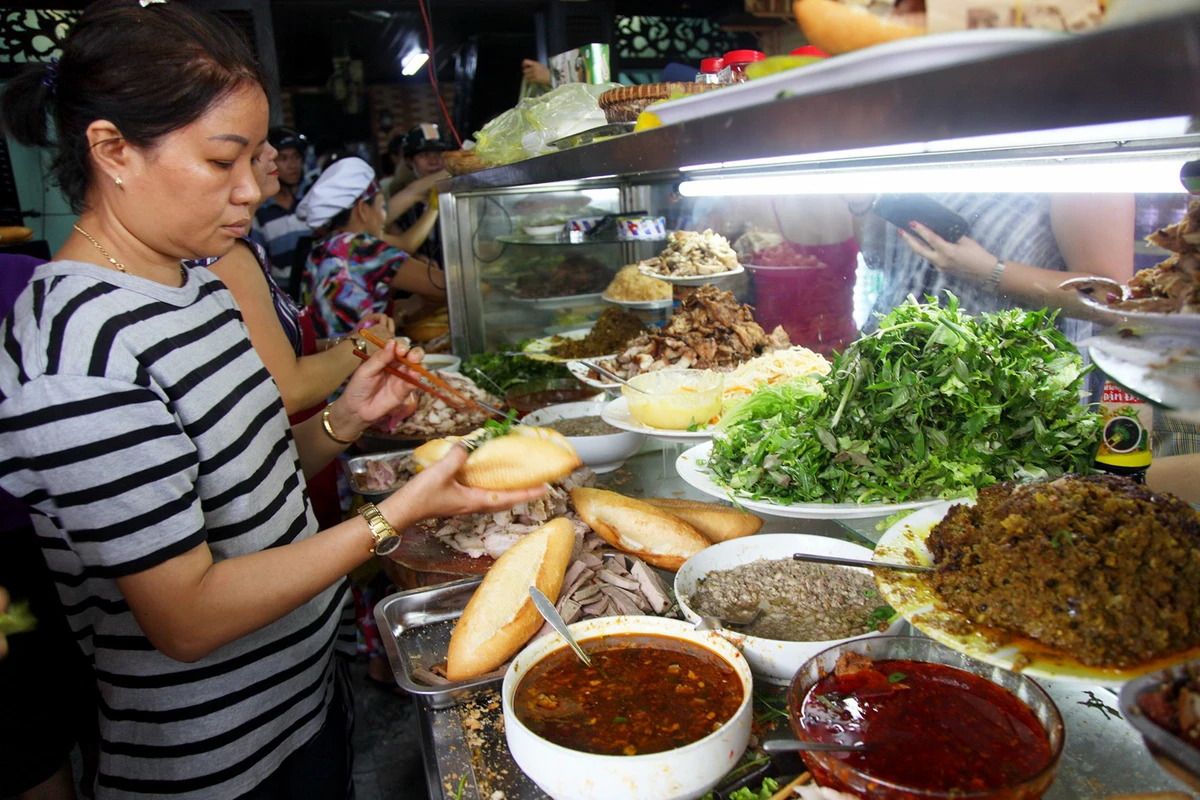 A woman prepares a Banh Mi sandwich at a busy Vietnamese street food stall filled with ingredients