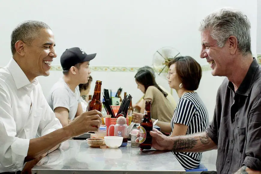 Barack Obama and Anthony Bourdain smiling and sharing beers while seated across from each other at a table in a casual eater
