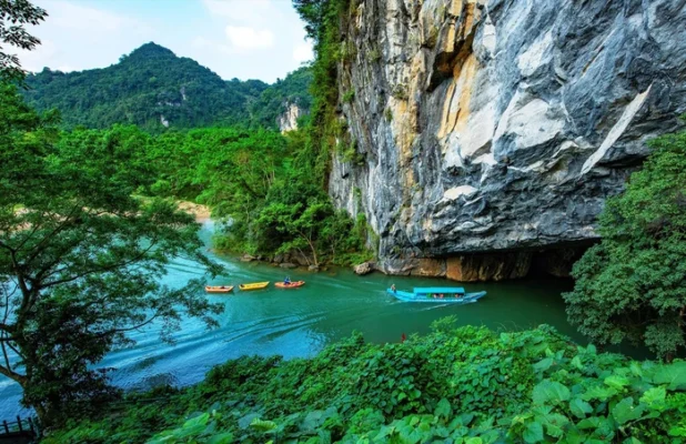 Boats on river by towering limestone cliff and lush greenery
