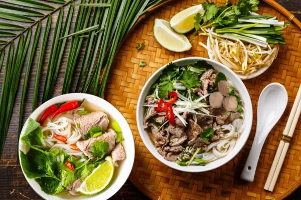 Two bowls of Vietnamese beef Pho with fresh herbs and lime on a bamboo tray