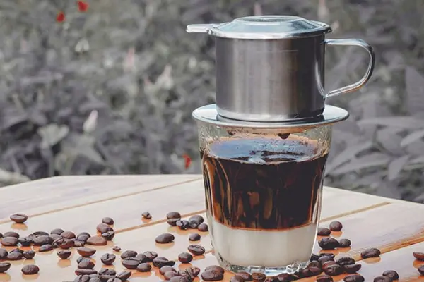Vietnamese phin coffee filter brewing into a glass with condensed milk, surrounded by coffee beans on a wooden table